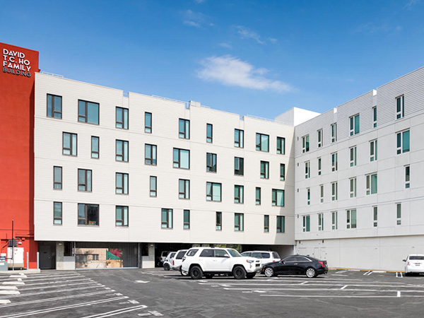 Photograph of a five-story, L-shaped apartment building in a modern style, taken from across an adjacent asphalt-topped parking lot.